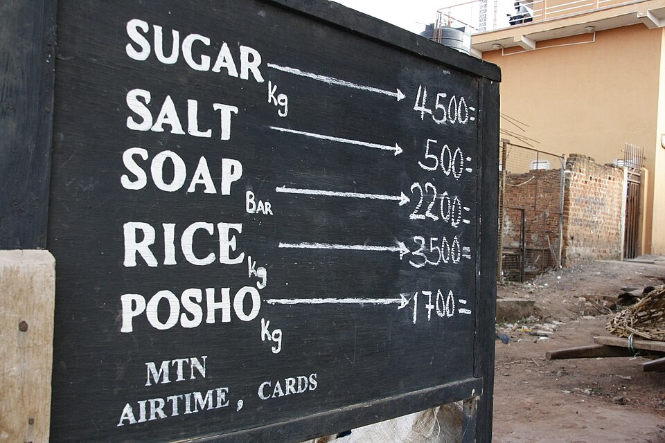 A hand-lettered board at a Ugandan market listing prices in shillings for sugar, salt, soap, rice and posho.