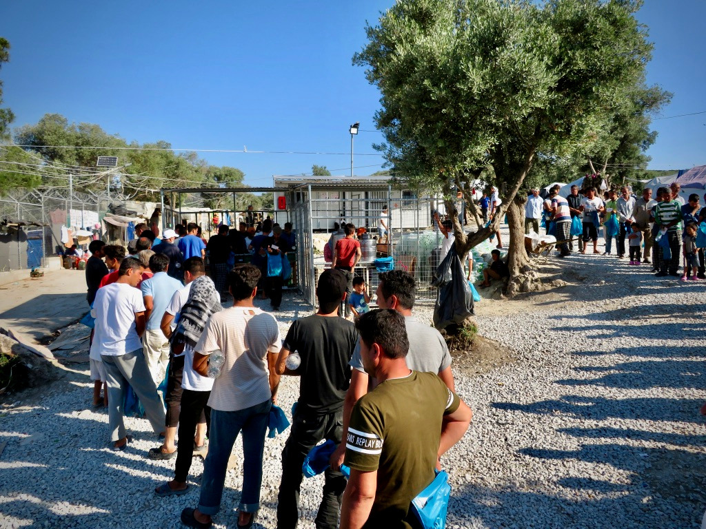 A queue of people waiting for a food distribution beside container units at Moria Camp.